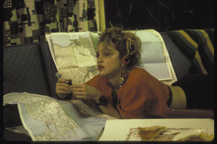 A still from Desperately Seeking Susan. Madonna is lying on her stomach on a couch while looking at a map and holding a pink telephone.